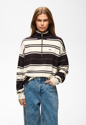 Ung kvinde iført en sort-hvid stribet lynlåstrøje med blå højtaljede jeans, stående foran en ensfarvet baggrund.