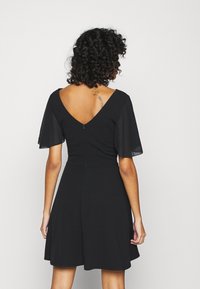 Zwarte jurk met een uitlopende rok, korte doorschijnende mouwen en een diepe V-hals aan de achterkant. Gladde stof met een verborgen rits aan de achterkant.