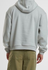 Man i ljusgrå hoodie och olivgröna byxor, sedd bakifrån mot en neutral bakgrund.