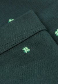 Tmavě zelená látka s hladkým povrchem a drobnými, světle zelenými květinovými vzory. Švy jsou viditelné podél okrajů.