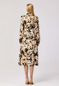 Robe florale mi-longue en beige avec des fleurs blanches et noires. Présente une taille cintrée, des manches longues et une texture lisse et douce.