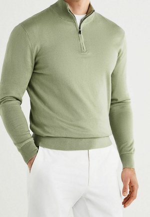 Homme portant un pull vert clair à fermeture éclair quart de longueur avec poignets et ourlet côtelés, associé à un pantalon blanc, debout avec une main dans la poche.