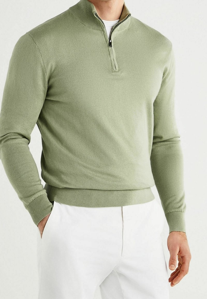 Homme portant un pull vert clair à fermeture éclair quart de longueur avec poignets et ourlet côtelés, associé à un pantalon blanc, debout avec une main dans la poche.