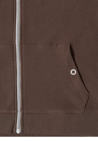 Gros plan sur la fermeture éclair d'une veste en tissu côtelé marron et une poche inclinée avec un petit détail métallique circulaire sur le bord de la poche.