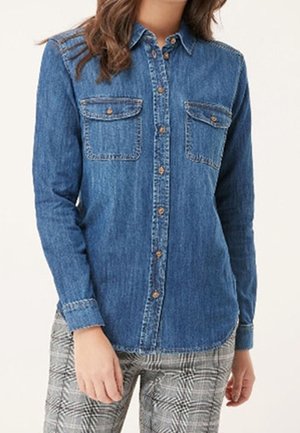 Femme portant une chemise boutonnée en denim bleu à manches longues avec deux poches poitrine et un pantalon à carreaux.