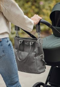 Šedá strukturovaná taška na rameno s dvojitými držadly a odnímatelným ramenním popruhem, vybavená přední kapsou a stříbrnými kovovými doplňky, držená osobou poblíž kočárku.