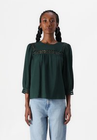 Top verde scuro con maniche a sbuffo, dettagli in pizzo sul petto e polsini a fantasia. Indossato con jeans azzurri chiari. Tessuto morbido e testurizzato.