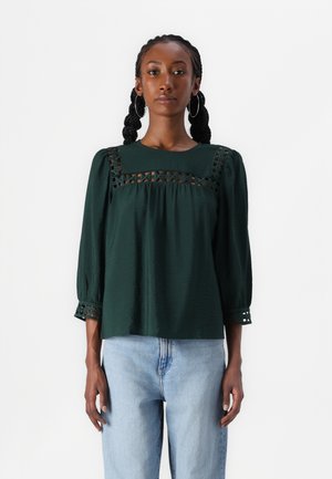 Mørkegrøn top med pufærmer, blonde detaljer over brystet og mønstrede manchetter. Båret med lyseblå jeans. Blødt tekstureret stof.