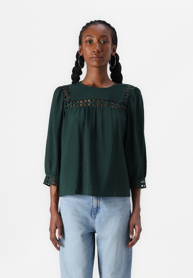 Top verde scuro con maniche a sbuffo, dettagli in pizzo sul petto e polsini a fantasia. Indossato con jeans azzurri chiari. Tessuto morbido e testurizzato.