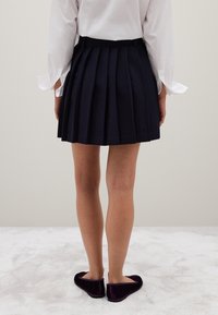 Falda de tablas navy con una cinturilla plana, combinada con zapatos de terciopelo morado tipo slip-on, vista desde la parte trasera, contra un fondo neutro.