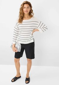 Haut à manches longues blanc avec des rayures noires, shorts noirs et sandales noires. Tenant un sac à main en métal argenté. Design simple et look décontracté.