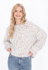 Pulover alb din tricot, cu pete colorate, având o croială lejeră, guler și tiv cu dantelă, și o textură pufoasă. Asortat cu blugi albaștri deschiși.