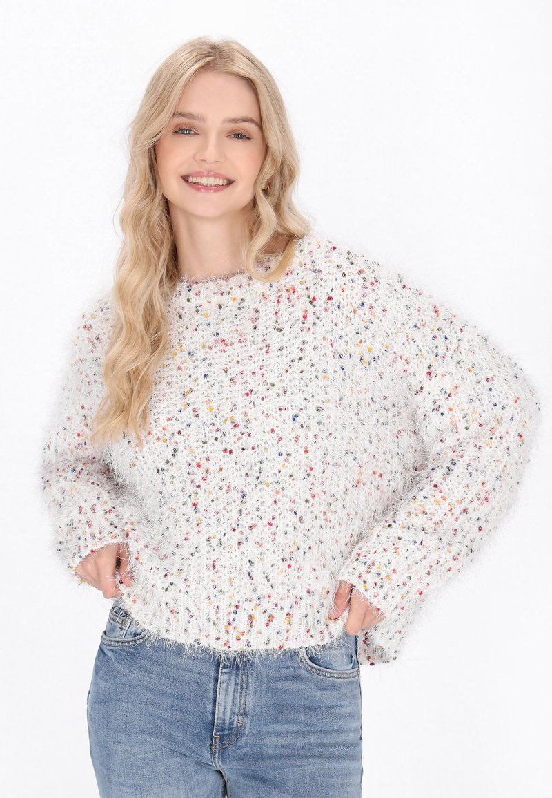 Pulover alb din tricot, cu pete colorate, având o croială lejeră, guler și tiv cu dantelă, și o textură pufoasă. Asortat cu blugi albaștri deschiși.