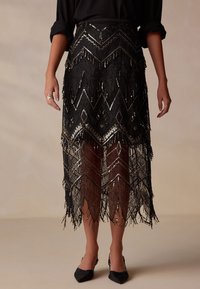 Black fringed skirt with geometric patterns, adorned with metallic accents and sequins, features a semi-sheer texture and scalloped hem.