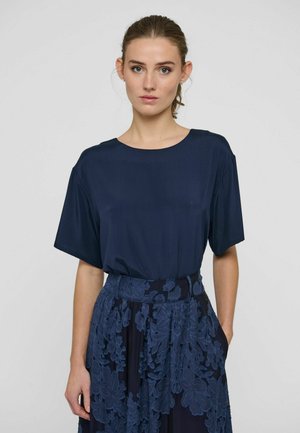 Femme à la peau claire et aux cheveux bruns attachés, portant un haut uni bleu marine à manches courtes et une jupe bleu marine ornée de détails en dentelle fleurie.