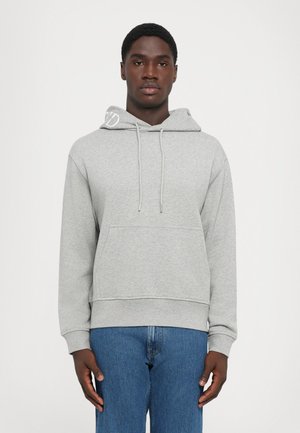 Jonge man in een lichtgrijze hoodie met een front pocket en een blauwe spijkerbroek, staande tegen een effen witte achtergrond.