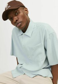 Camisa de manga corta azul claro con cuello y un bolsillo en el pecho. Tela suave con cierre de botones visible. Lleva una gorra marrón.