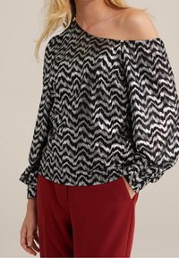 Černá a stříbrná halenka se vzorem cikcak, s odhalenými rameny, dlouhými nafouklými rukávy a elastickými manžetami. Kombinováno s červenými kalhotami.