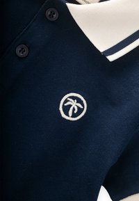 Polo shirt blu navy con colletto a contrasto crema, caratterizzato da ricami bianchi di palme e bottoni scuri. Tessuto a trama, design casual.