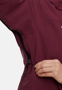 Vinkolorerad, texturerad tygjacka med dragkedja och snörning vid midjan, med en närbild av en hand som justerar snörningen.