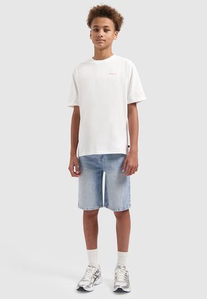 Jugendlicher Junge steht, trägt ein weißes kurzärmeliges T-Shirt, hellblaue Jeansshorts, weiße Socken und silberne Sportschuhe.