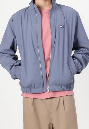 Person trägt eine hellblaue Jacke mit Reißverschluss über einem pinken T-Shirt und beigen Hosen, die Hände in den Jackentaschen.