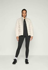 Puffjakke i lys beige, med quiltet design, frontlås, kombinert med en tettsittende svart topp, svarte leggings og svarte sneakers.