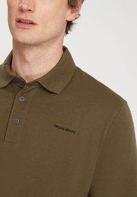 Oliwkowa koszulka polo z kołnierzykiem, dwoma guzikami i teksturowanym materiałem. Posiada małe haftowane logo na lewej piersi.