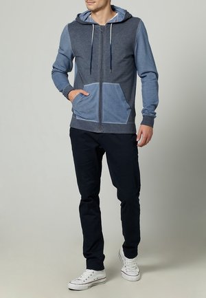 Felpa blu con zip e design bicolore, dotata di tasca frontale e cappuccio regolabile. Abbinata a pantaloni scuri e sneakers bianche.