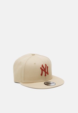 Beige Flachkrempe-Baseballkappe mit rotem, gesticktem NY-Logo auf der Vorderseite und einem runden schwarzen und goldenen Aufkleber auf der Krempe.