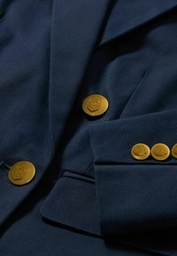 Námořnický blejzr s texturovanou tkaninou, zdobený zlatými knoflíky a přiléhavým střihem. Významné detaily zahrnují designy erbu na knoflících.