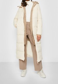 Lang beige puffet frakke med hætte, der har en lynlåslukning i front, en blød brun indvendig foring og sidelommer. Sammen med beige bukser og sneakers.