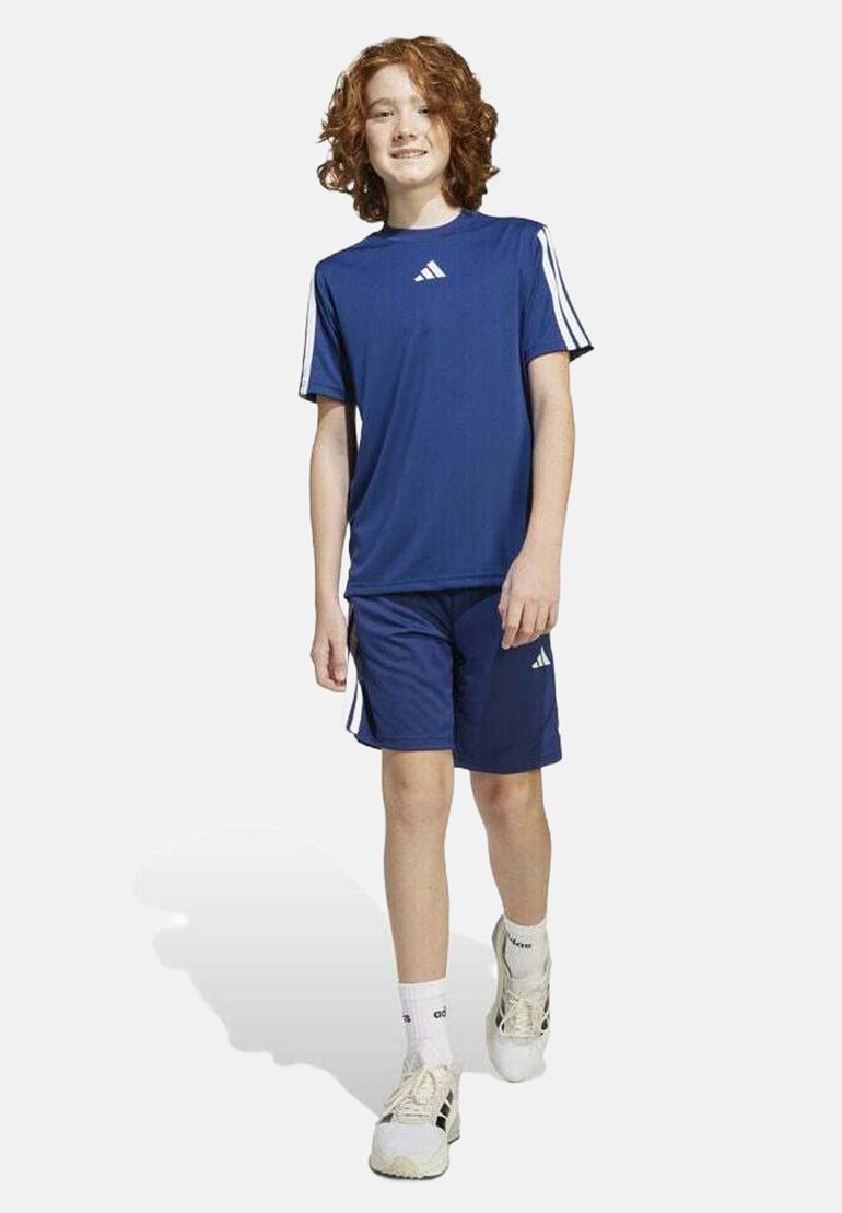 Camiseta atlética azul con rayas blancas en las mangas, combinada con pantalones cortos azules a juego. Llevado con calcetines blancos y zapatillas deportivas ligeras.
