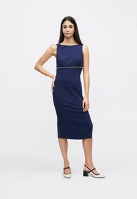 Μίντι φόρεμα σε ναυτικό μπλε χωρίς μανίκια, με εφαρμοστή σιλουέτα και διακοσμητικό με χάντρες στη μέση· ομαλή υφή. Το μοντέλο φορέθηκε με μαύρα και λευκά ψηλοτάκουνα παπούτσια.