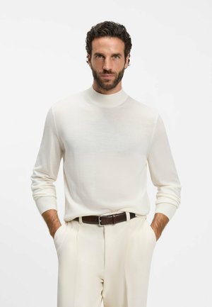 Uomo con capelli corti, scuri e ricci e barba, che indossa un maglione bianco a collo alto, pantaloni beige e una cintura marrone, con le mani nelle tasche.