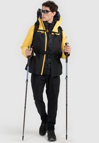 Homme portant des lunettes de soleil, une veste d'extérieur jaune et noire, un pantalon noir et des chaussures de randonnée, tenant des bâtons de marche, marchant vers l'avant.