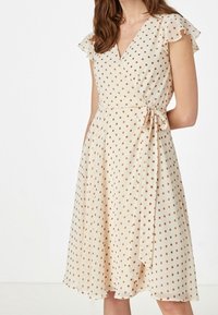 Krämfärgad omlottklänning med korta, volangdetaljerade ärmar, prydd med guldprickar, midjeband och en svängd kjol som faller under knäet.