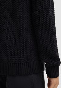 Ciemny sweter z dzianiny z teksturowanym wzorem, ściągaczami przy mankietach i dolnym brzegu. Materiał wydaje się miękki, z luźnym krojem i szerokimi rękawami.