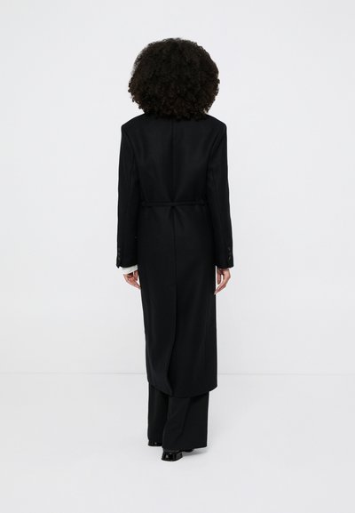 Must pikk mantel, millel on istuv vöökoht, tagumine vöödetail ja kohandatud varrukad. Sile tekstuur ja struktureeritud disain, kombineeritud laia säärega pükstega.