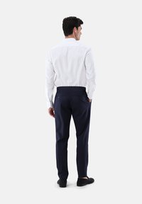 Weißes Langarmhemd, marineblaue Hose, glatter Stoff, taillierte Passform, doppelte Gesäßtaschen mit Knöpfen, getragen mit schwarzen Slippern.