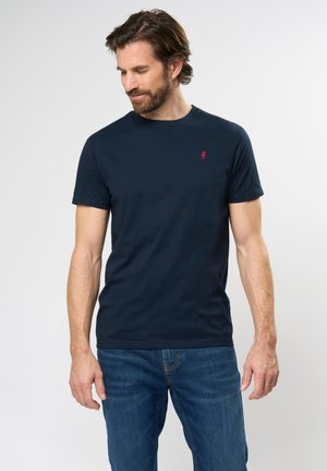 Marineblå crewneck t-skjorte laget av bomull, med en liten rød logo på venstre bryst. Kombinert med blå denimjeans.