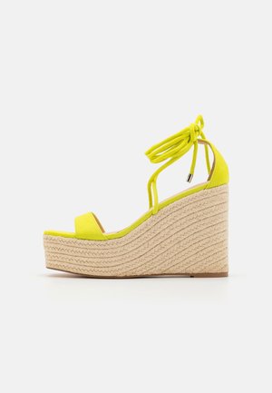 Sandale compensée jaune avec semelle plateforme en corde tressée et brides à nouer à la cheville sur fond blanc.