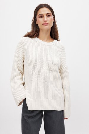 Pull en mailles crème avec texture côtelée, encolure ronde et manches tombantes, associé à un pantalon gris foncé.