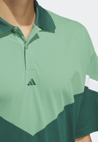 Mies, der ein grün und dunkelgrün blockiertes Polohemd mit einem dunkelgrünen Kragen und dem Adidas-Logo auf der Brust trägt.