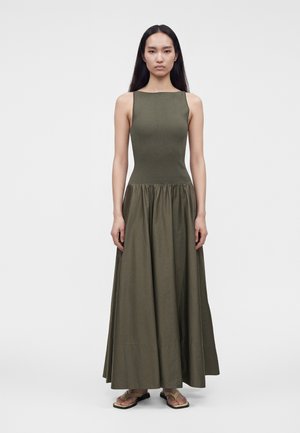Γυναίκα με μακριά μαύρα μαλλιά φοράει αμάνικο ελιάς πράσινο φόρεμα με εφαρμοστή κορσέ και φούστα σε γραμμή Α, στέκεται μπροστά σε απλό λευκό φόντο.