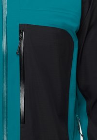 Teal en zwarte jas van een textuurstof, met een voorzak met rits en trekkoordaccenten aan de onderkant voor aanpassing.