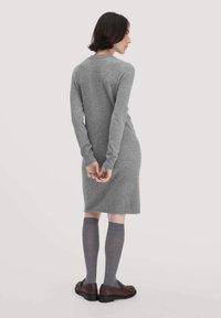 Donna in piedi con le spalle rivolte, che indossa un abito lungo a maniche lunghe grigio, calzini grigi fino al ginocchio e mocassini marroni su uno sfondo chiaro.
