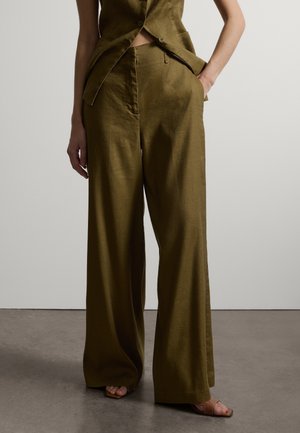 Person trägt weite, olivgrüne Hosen mit weitem Bein und eine passende ärmellose Weste, steht mit einer Hand in der Tasche.