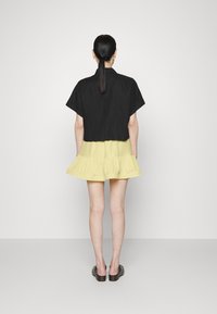 Top preto cortado com mangas curtas combinado com um vestido amarelo com babados até ao joelho. Modelo a usar sandálias de salto pretas, vista de costas.