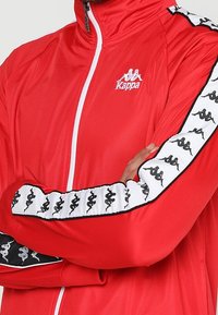 Rote Zip-Up-Jacke aus leichtem Stoff. Mit weißen Akzenten und einem schwarz-weiß gemusterten Logo-Design entlang der Ärmel.
